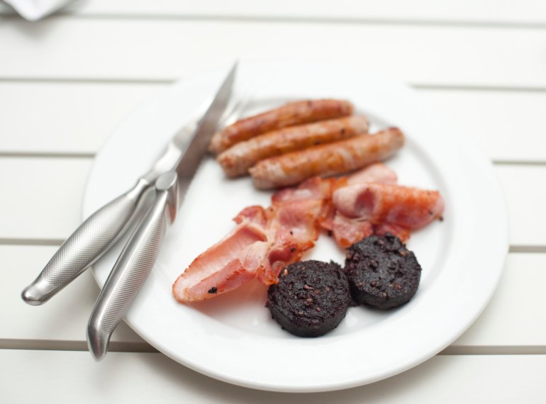 Fried breakfast with mushrooms, crispy bacon and pork sausages served on a white plate on a painted white slatted table with shallow dof