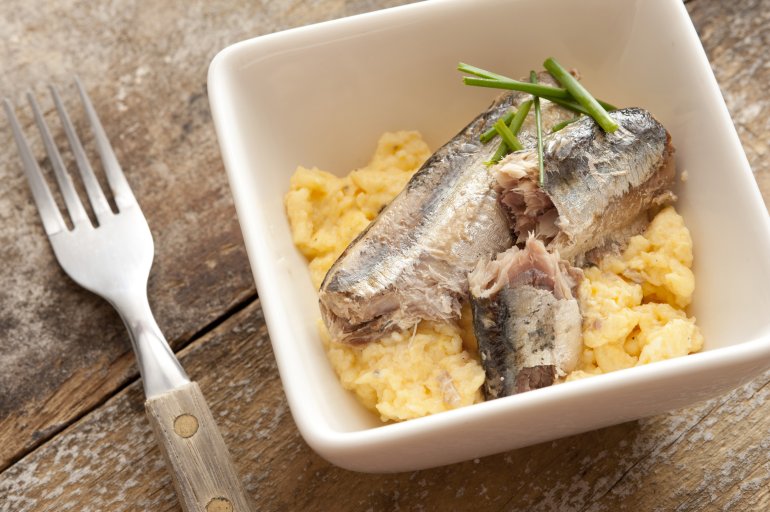 Delicious breakfast of sardines with chives and scrambled eggs in little square bowl beside fork on old wood surface