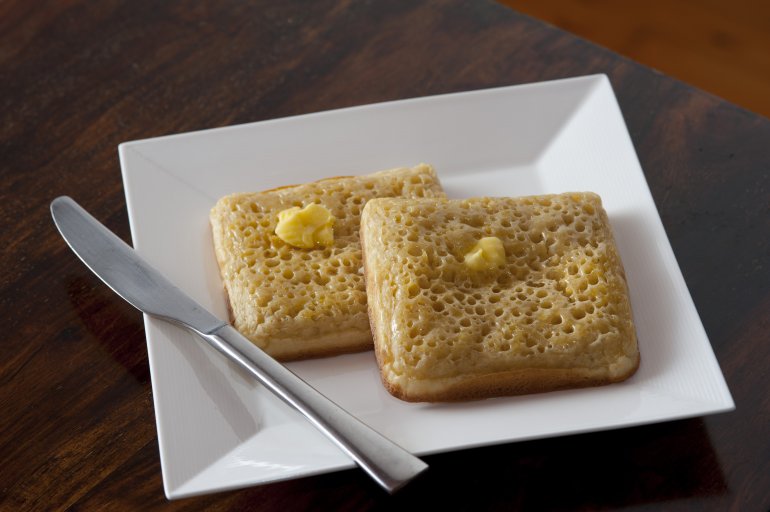 Two hot buttered square shaped crumpets on a modern square white plate with a knife on the side