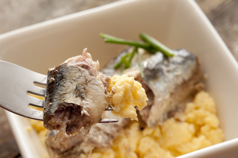 Close up of sardines on fork above delicious scrambled egg breakfast garnished with green garlic chives