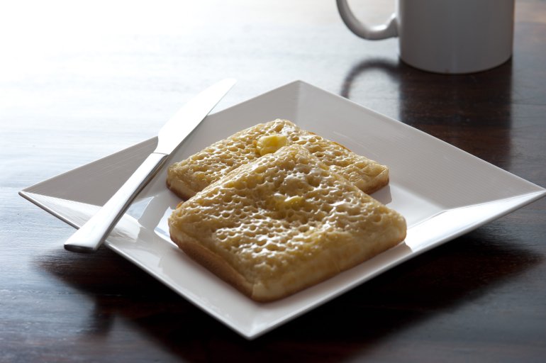 Modern breakfast of two square crumpets with knife on a white plate besides a coffee mug