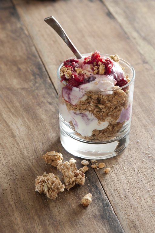 Little glass cup of granola muesli cereal with yogurt and spoon inside on wooden table beside crumbs