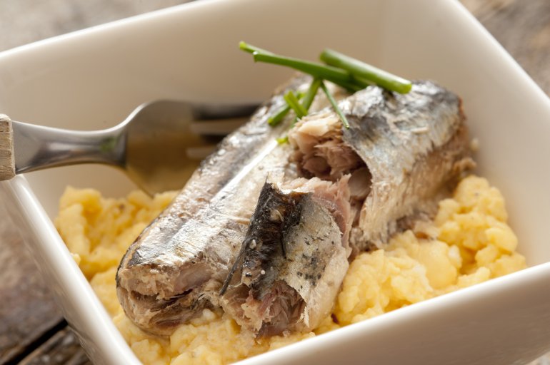 Close up view on fork placed in sardine breakfast surrounded by scrambled eggs and garnished with garlic chives in square bowl
