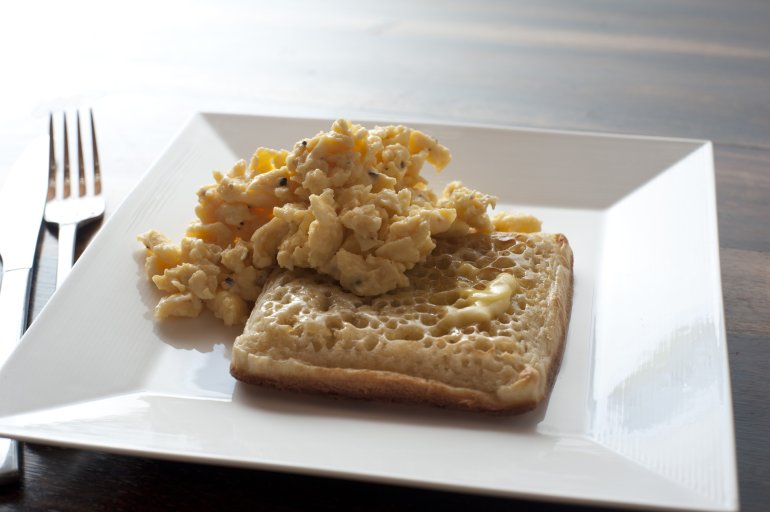 Single delicious buttered square crumpet and scrambled eggs in plate next to fork and knife on table