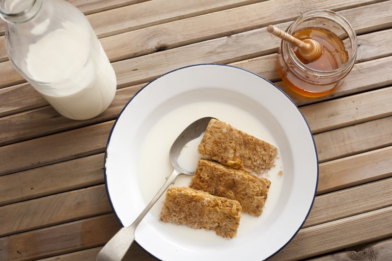 First person top down perspective view on whole wheat breakfast cereal bars in round bowl with honey and bottle of milk over table