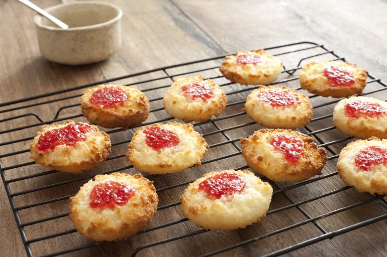 Oven tray full of baked coconut cookies with strawberry jam in the middle over table next to dish