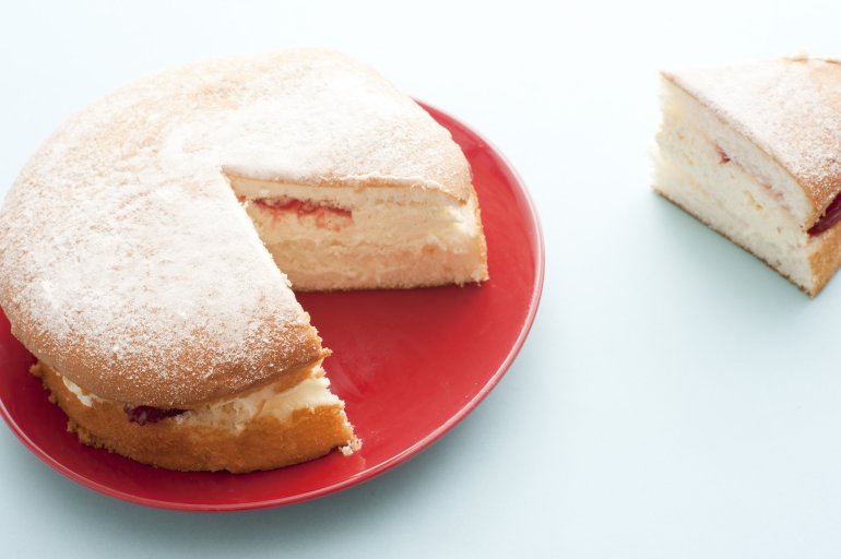 Freshly baked simple sponge cake sprinkled with icing sugar and filled with cream and strawberry jam with one slice cut and moved to the side of the plate