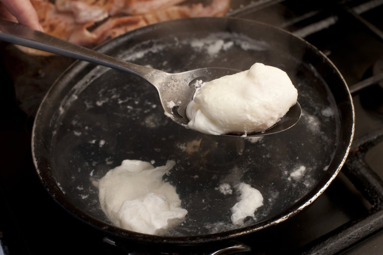 Person poaching eggs in boiling water in a frying pan over a gas hob lifting one clear with a spoon