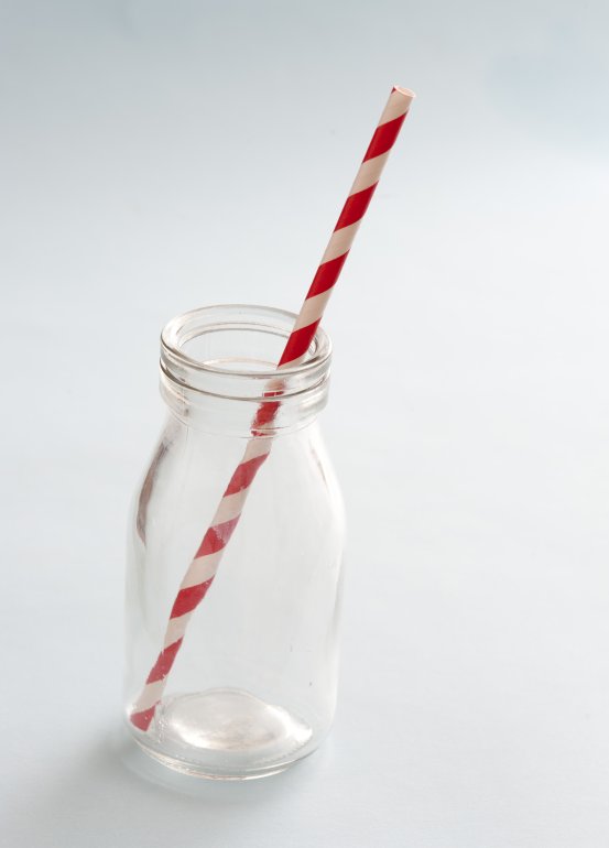 Empty kids glass milk bottle with striped red and white plastic straw over a white background with copy space
