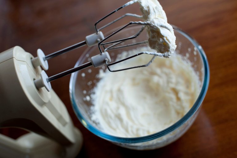 Electric kitchen mixer with a metal whisk attachment coated in thick whipped cream from the glass mixing bowl below