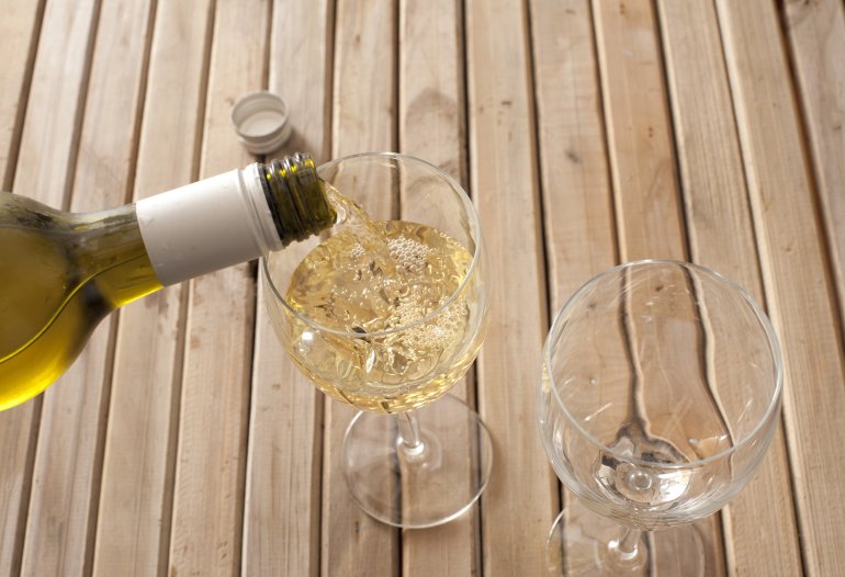 Pouring a glass of white wine from a bottle into a wineglass standing on a slatted wooden table, close up high angle view of the neck of the bottle and glass, with copyspace