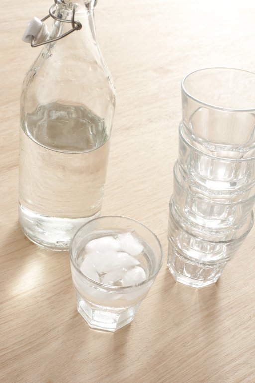 Glass bottle of pure fresh water standing on a wooden table alongside a stack of tumblers and glass filled with liquid