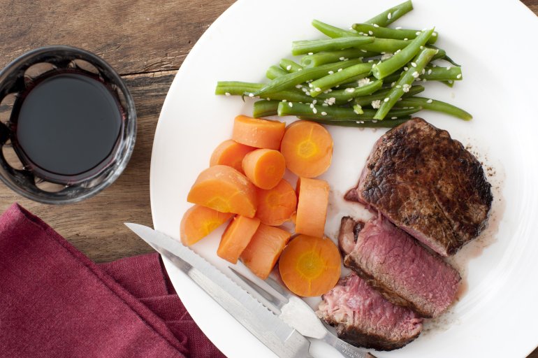 Sliced grilled fillet steak and cooked vegetables with diced carrots and savory green runner beans served with a glass of red wine