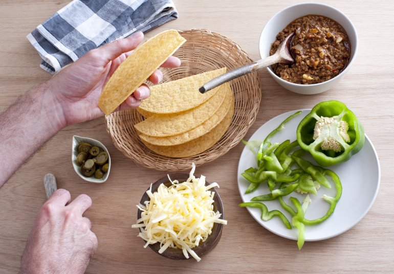 High Angle View of Male Hands Preparing Tacos Using Freshly Prepared Ingredients - Seasoned Beef, Green Pepper, Shredded Cheese, Jalapenos and Hard Corn Shells