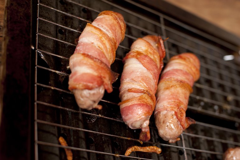 Three bacon sausage rolls being prepared on grill, viewed in close-up