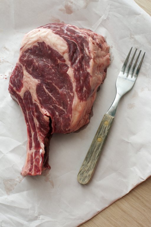 Bone-in thick juicy raw ribeye beef steak on crumpled white butchers paper with fork