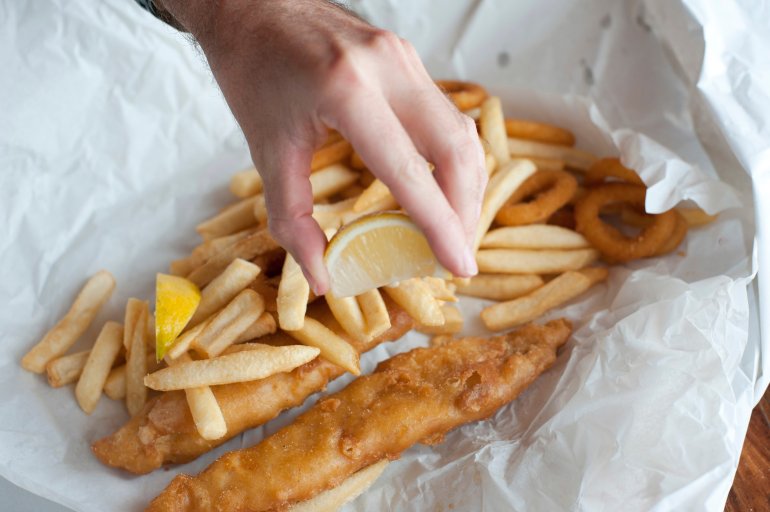 Hand squeezing lemon over takeaway fried fish fillets and calamari in batter with fried potato chips on crumpled paper