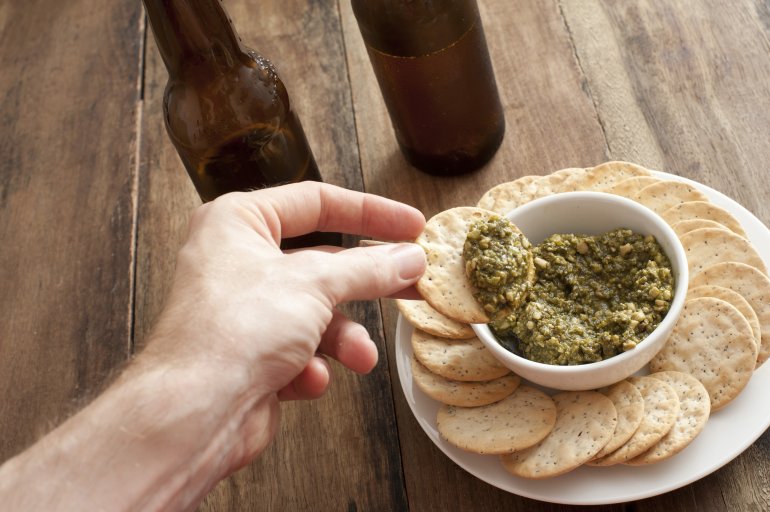 Man dipping a cracker in a savory dip in a bowl on a plate with two bottles of beer alongside on a wooden counter