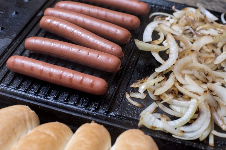 Preparing a tasty hot dog on a grill with smoked Wiener sausages, diced onions and fresh white bread rolls