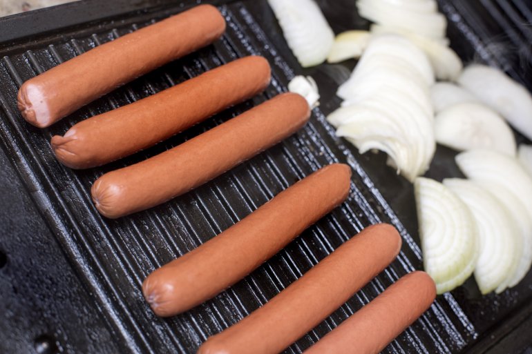 Close up on six hot dogs and various chopped white onions cooking on large black grill