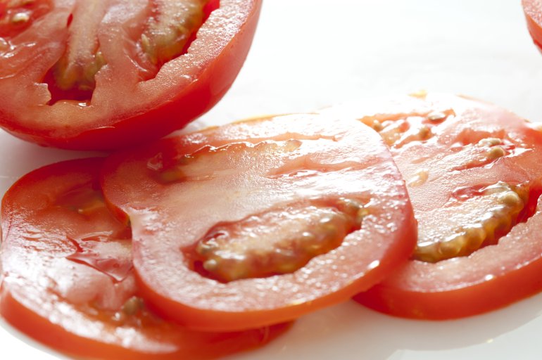 Slices of fresh ripe red tomato lying on a white background in front of a halved juicy tomato, close up view