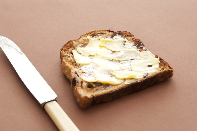 Buttered toasted bread with a thick spread of butter on a slice of fruity raisin bread on a brown background with a knife