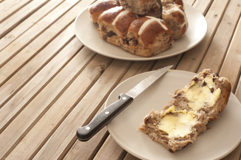 Buttered Easter Hot Cross Bun with fruity raisins served on a side plate ready for a tasty refreshment at teatime, wooden slatted tale with copyspace