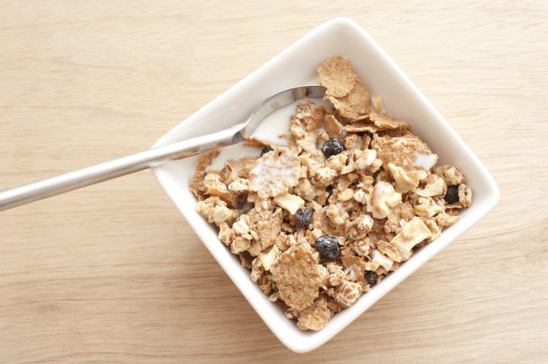 wheat and granola cereal First person perspective view of spoon in square bowl of muesli granola cereal with slices of dried fruit on top with milk over wooden table