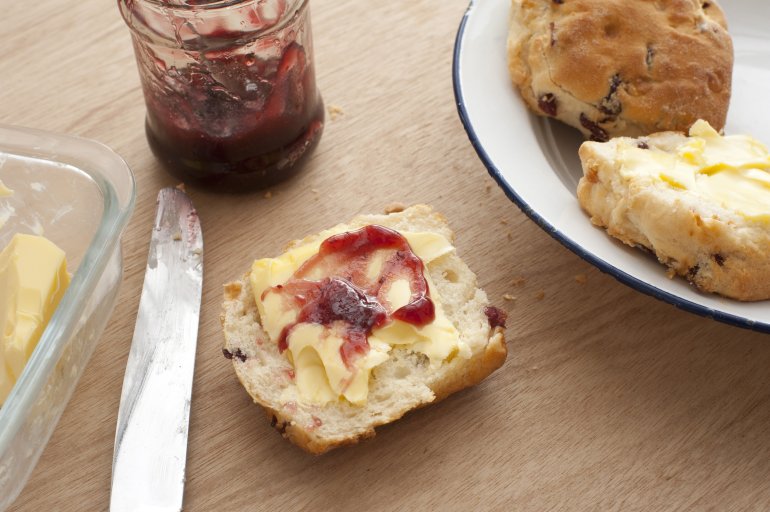Half buttered scone with strawberry jam on a wooden table alongside the jam jar, plate of scones and pat of butter