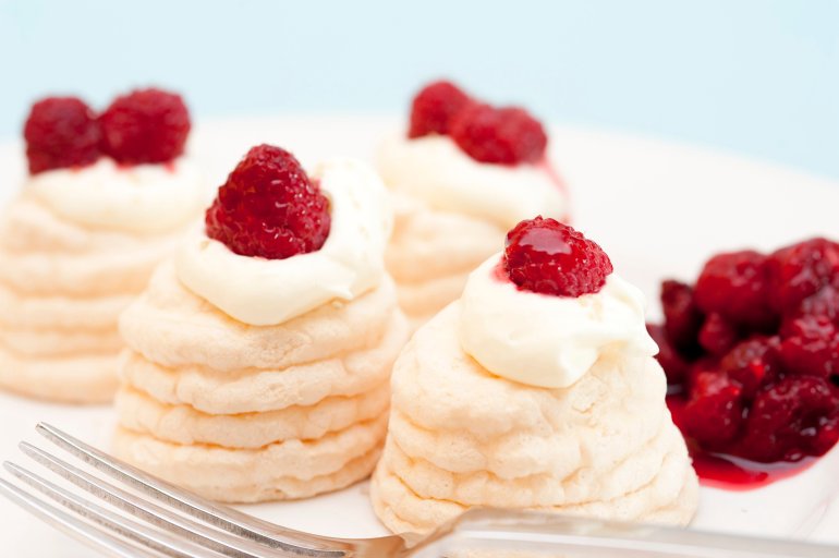 Individual twirled meringue cases filled with whipped cream and topped with fresh raspberry compote served on a plate