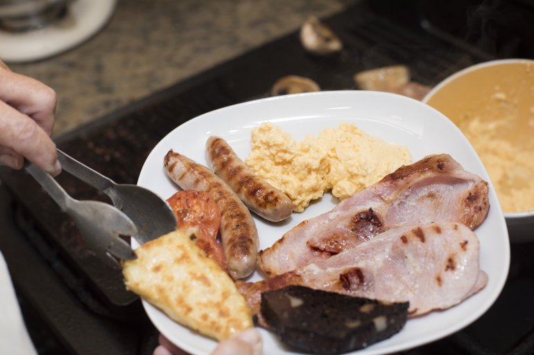 Hand using stainless steel tongs to place a hash brown beside sausage and ham on breakfast plate