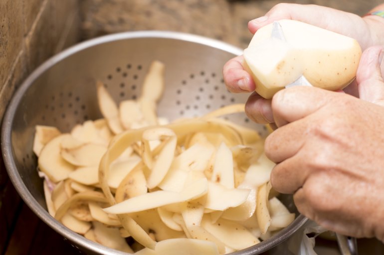 Man peeling fresh potatoes for cooking catching the peels in a metal colander, close up on his hands and the utensil