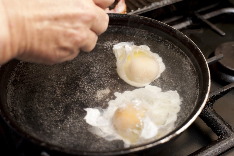 preparing two poached eggs Overhead view of hands preparing two eggs in boiling water set on a gas stove and an iron skillet