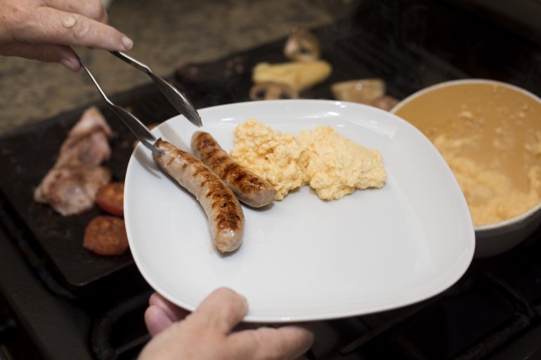Man preparing hot breakfast Man preparing hid breakfast serving up to cooked sausages from a grill onto a modern white plate, close up of his hands