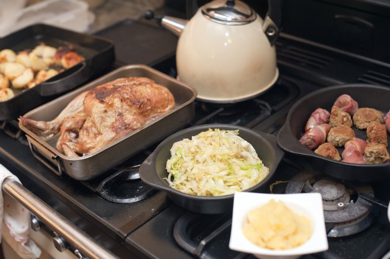 Roasted chicken dinner preparation Roasted chicken sits in pan beside tea kettle, sausage wrapped in bacon and other cooked foods all resting on a gas range