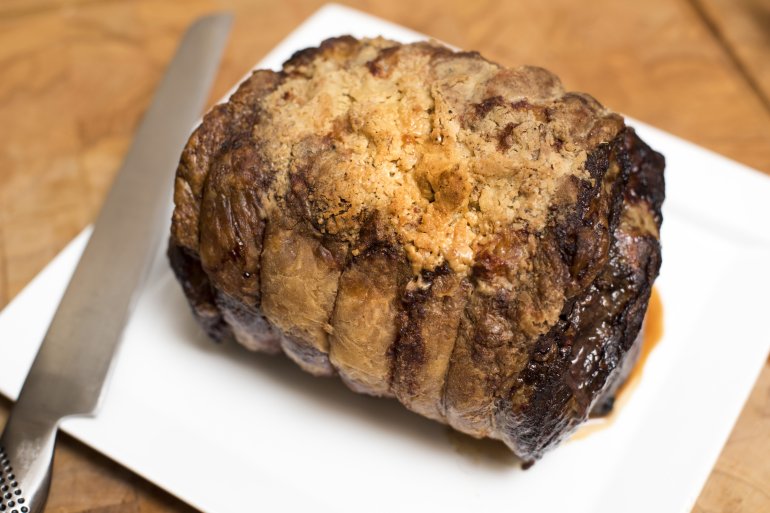 Overhead view of roast beef portion tied with string and sitting on white cutting board beside sharp cleaver