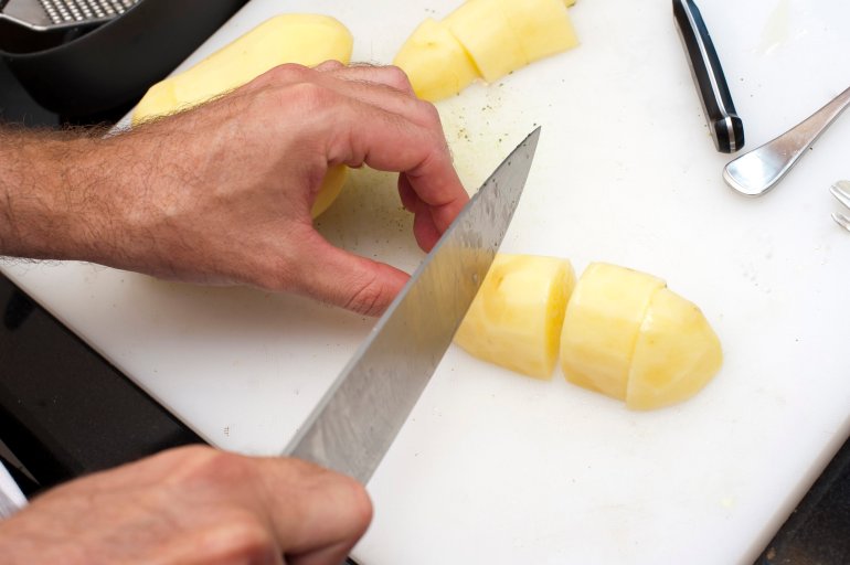 Man cutting peeled potatoes on a chopping board in the kitchen with a large chefs knife as he prepares the vegetables for the meal