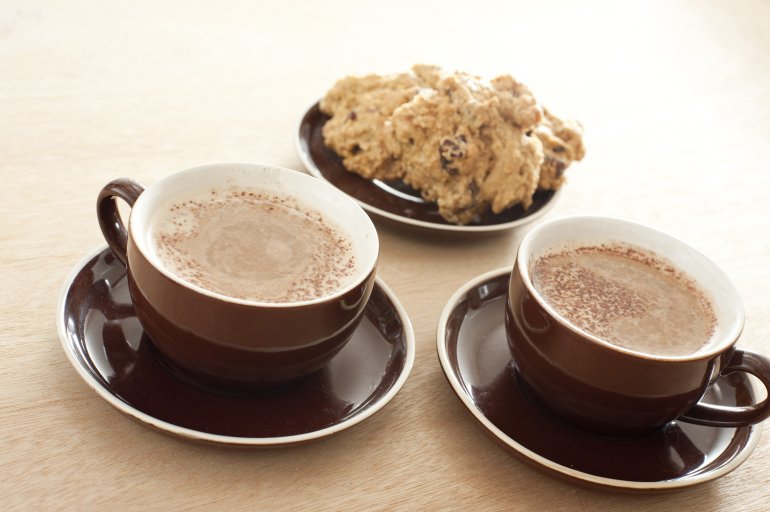 Two cups of coffee with cake Two brown ceramic cups and saucers of fresh frothy coffee served with a small plate of sliced cake on a graduated beige background