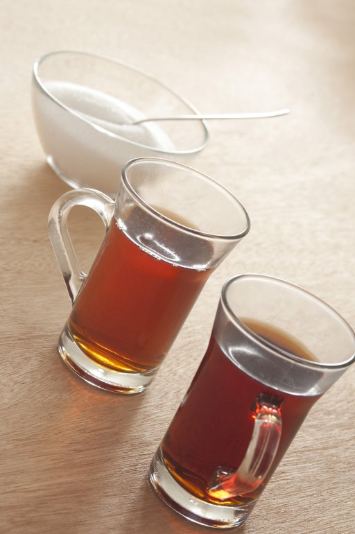 Two glass mugs of freshly brewed hot black tea with a bowl of sugar behind on a yellow background, tilted high angle view