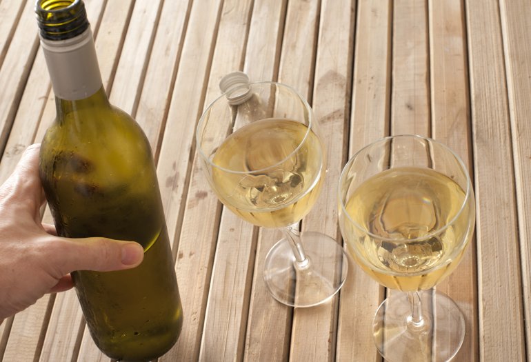 Man pouring white wine from a bottle into two glasses on a wooden table, close up high angle view of his hand with copyspace