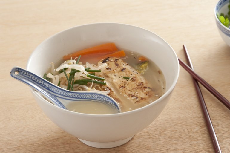 Single white bowl of Asian noodle soup filled with broth, carrots and other vegetables next to chopsticks