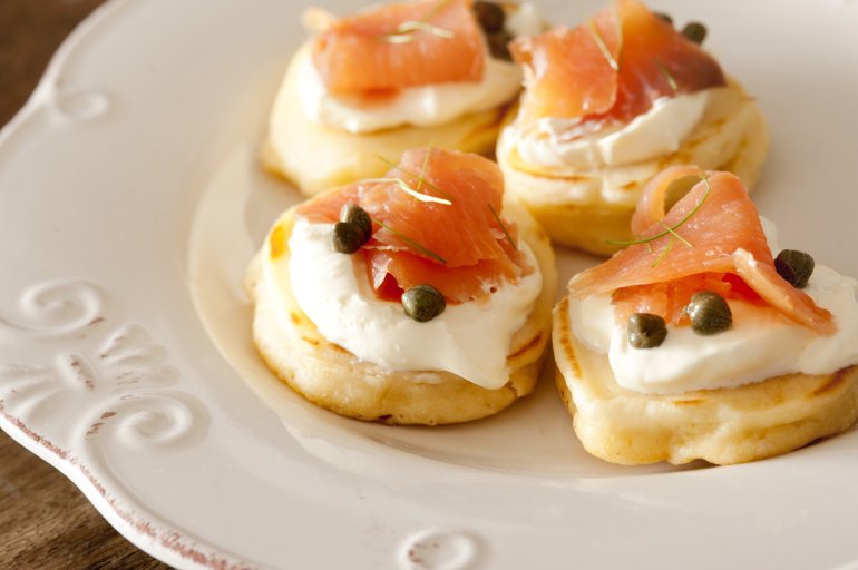 Four raw fish and cheese blini snacks with capers and herbs on white glossy ceramic plate