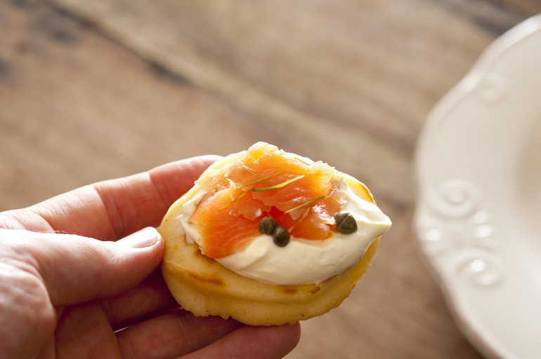 First person view on hand holding delicious blini snack topped with creme and pink salmon over wooden table