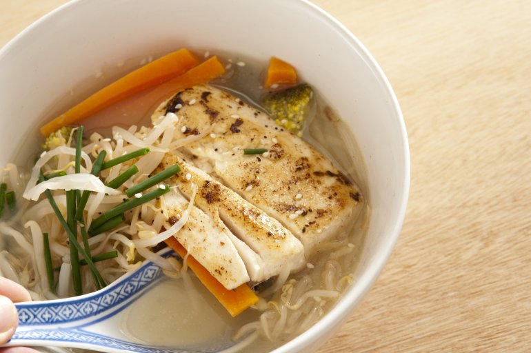 Hand holding a decorative blue and white spoon in milkfish soup bowl with noodles, chives and carrots in the broth