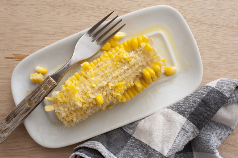 Eaten sweet corn cob with remnants of kernels and a fork on a white plate, view from above
